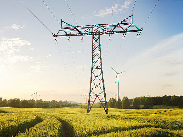 A power pole stands in a field, with a wind turbine visible on the horizon.