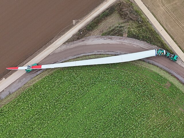 The blade of a wind turbine is being transported along a dirt track by two green lorries.