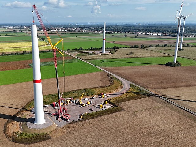 The tower of a wind turbine is being erected on a brownfield site. There are other fields and wind turbines in the surrounding area.
