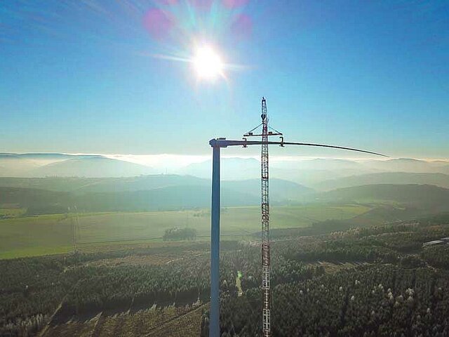 Ein Windrad steht in einem Waldgebiet neben einem Kran. Die Sonne scheint vor einem wolkenlosen, blauen Himmel.
